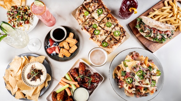 Assorted shareable food dishes arranged on a table, including flatbread pizza, chicken wings with dipping sauce, nachos, sandwiches with fries, and mixed drinks